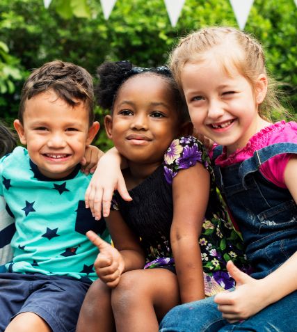 Three kids smiling together outdoors and giving thumbs up